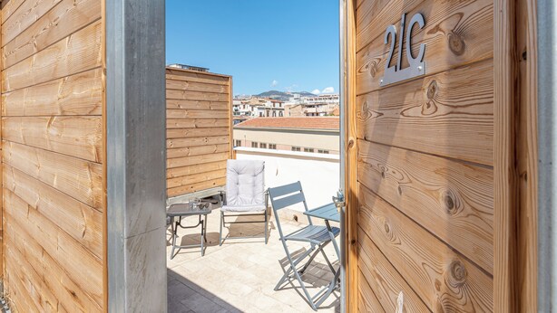 Pared exterior de madera con un letrero '2G', un balcón con una mesa y dos sillas, y vistas a edificios en el fondo.