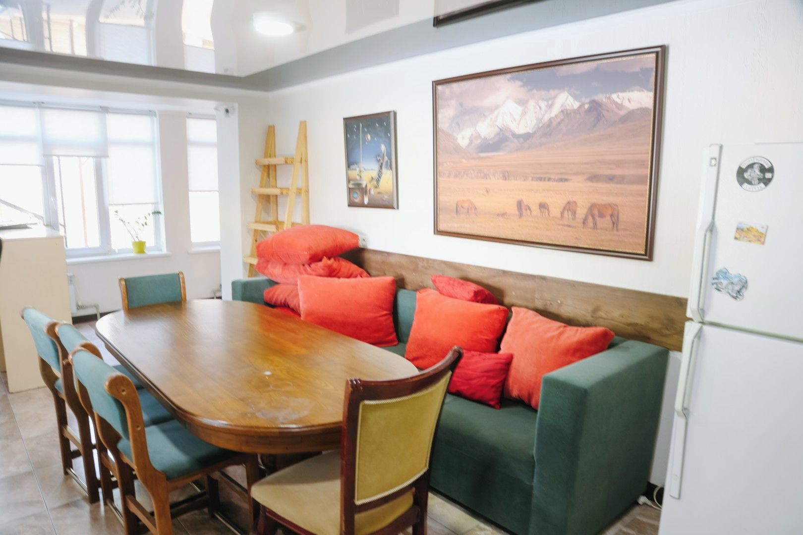 A dining area with a wooden table and chairs, a green sofa with red cushions, and a painting of a mountainous landscape.