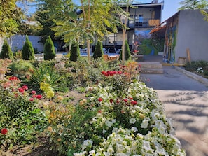 Un jardín con diversas plantas y flores, un edificio con mesas al aire libre y un mural en la pared.