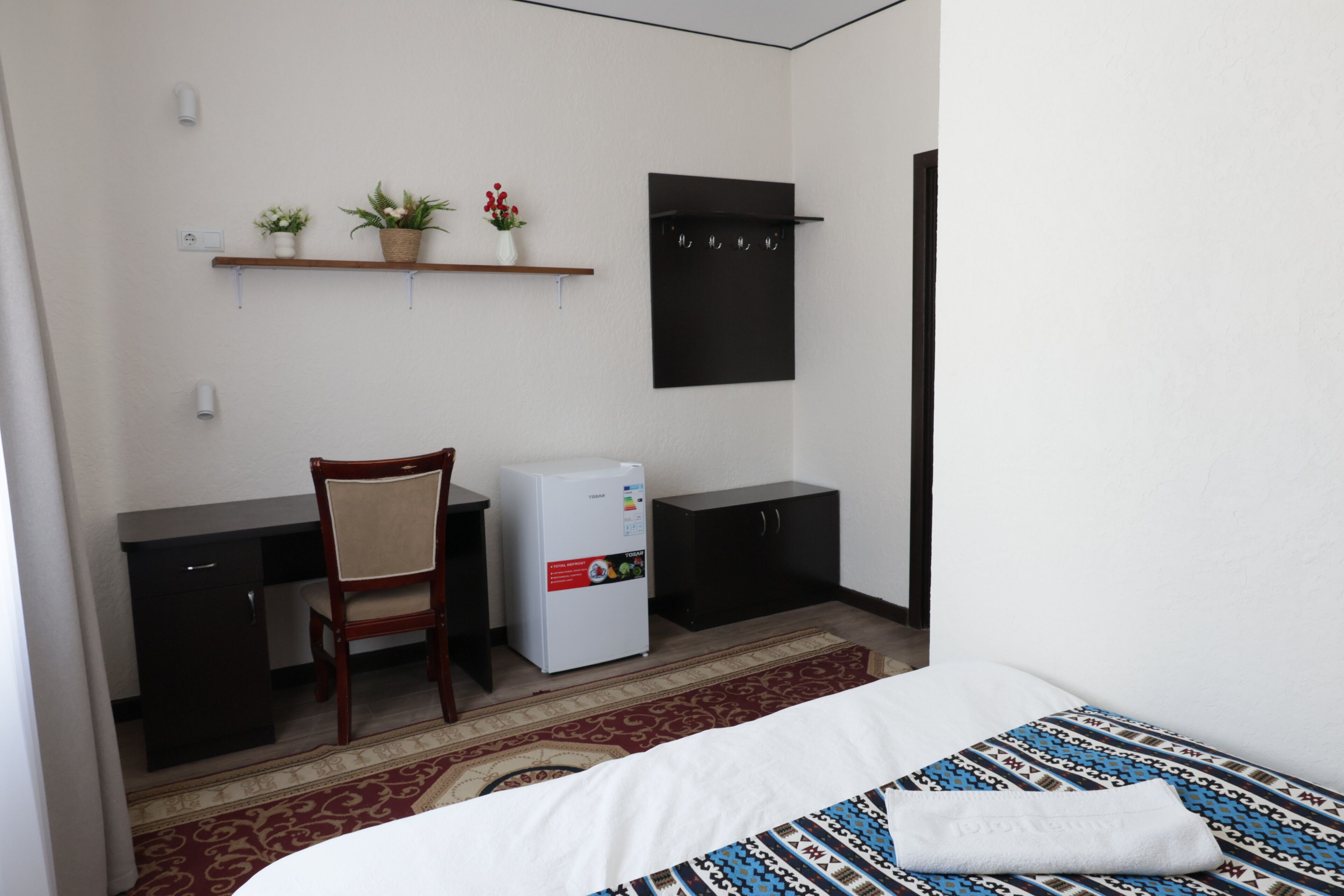 A hotel room with a bed, a desk, a chair, a refrigerator, a wardrobe, and a shelf with plants.