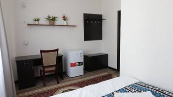 A hotel room with a bed, a desk, a chair, a refrigerator, a wardrobe, and a shelf with plants.