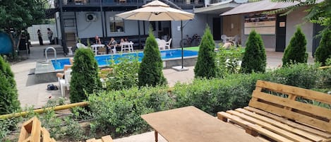 Zona de piscina exterior con muebles de madera, un edificio con balcones y plantas en macetas.