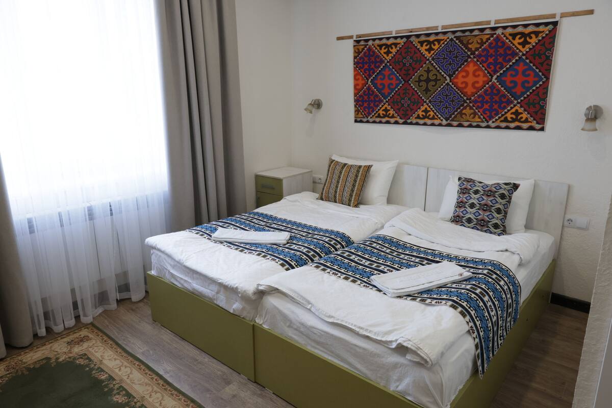 Un dormitorio con cama, mesita de noche, tapiz en la pared y ventana con cortinas.