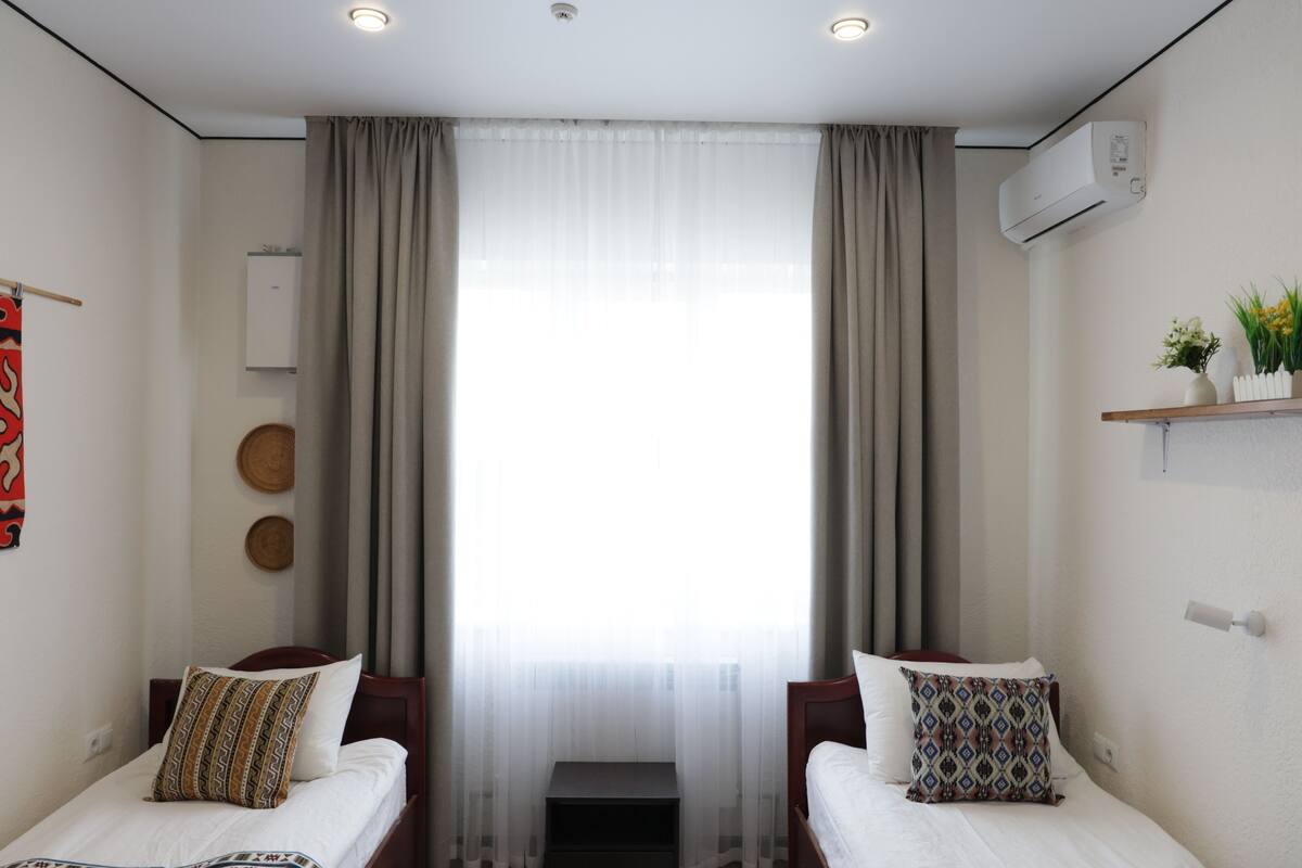 Habitación de hotel con dos camas, una ventana con cortinas, un equipo de aire acondicionado y un pequeño estante con adornos.