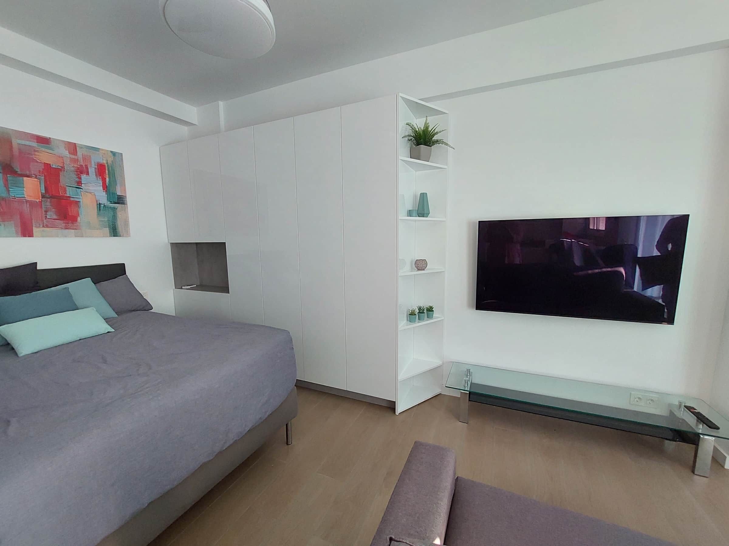 Moderni makuuhuone, jossa on sänky, televisio, lasinen sohvapöytä ja ruukussa oleva kasvi.