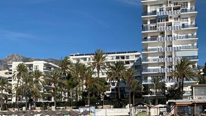 Un hotel de varios pisos con balcones, palmeras y montañas al fondo.