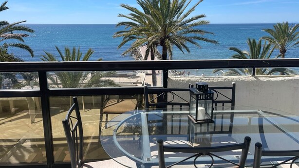 Un balcón con una mesa y sillas de vidrio, con vista a una playa con palmeras y el mar.