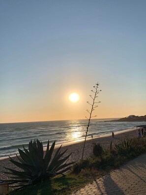 Ein Strand bei Sonnenuntergang über dem Meer, bewölkter Himmel und einige Personen, die am Ufer entlanggehen.