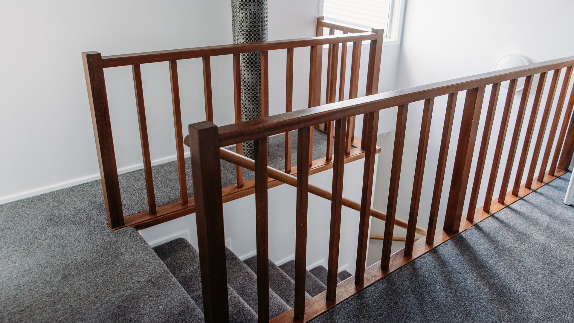 A wooden staircase with a handrail, carpeted steps, and a white wall in the background.