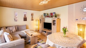 Un salon comprenant un canapé, un téléviseur, un meuble de rangement en bois et une table ronde ornée d’un vase de fleurs.