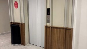 En hall med en soptunna, en dörr med ett vertikalt panel och en väggmonterad manöverpanel för hissen.