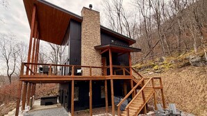 Una casa moderna con una terraza de madera, una chimenea de piedra y una escalera que lleva a un segundo nivel.