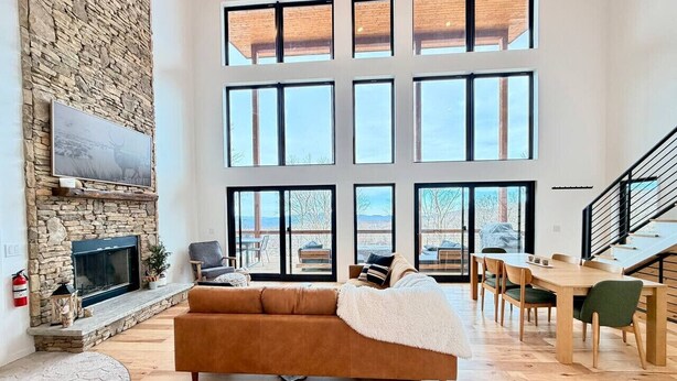 Amplia sala de estar con chimenea de piedra, ventanas grandes, un sofá y una mesa de comedor.