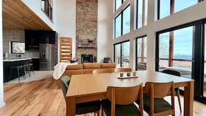 Una cocina moderna con chimenea de piedra, comedor con mesa y sillas de madera, y ventanas amplias con vistas.