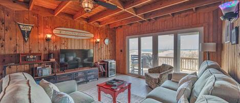 A cozy cabin interior with a large TV, wooden walls, a ceiling fan, and a view of the beach through the sliding glass door.