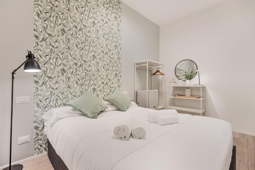Una cama bien hecha con almohadas, una lámpara de noche, un espejo y un estante con artículos de tocador.
