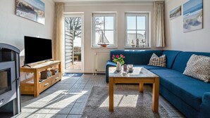 A living room with a blue sofa, a wooden coffee table, a flat-screen TV, and a window with a view of the outside. - Haus Boddenkieker 1 Rügen in Mönchgut – Ferienwohnung mit Meerblick und Südterrasse (Mönchgut)