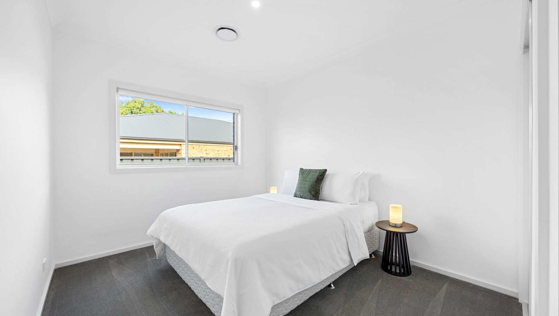 Une chambre à coucher avec un lit, une table de chevet avec une lampe, une fenêtre donnant sur des immeubles et un sol recouvert d’une moquette.