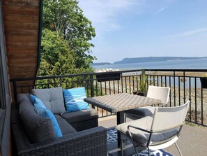 A balcony with a sofa, chairs, and a table, overlooking a body of water and hills.