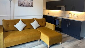 A modern living room with a mustard-colored sofa, a small ottoman, a kitchen with dark cabinets and a white countertop, and a painting of a pier on the wall. - Peaceful 1 bedroom house. Dog friendly, centrally located hidden gem. (Morpeth)