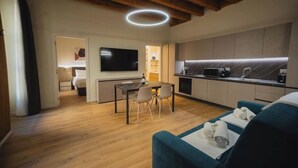 Habitación de hotel moderna con techo de madera, televisor de pantalla plana, zona de comedor con mesa y sillas, kitchenette y sofá azul con toallas.