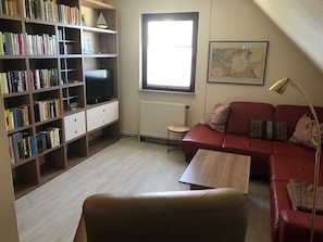 Een woonkamer met een boekenkast, een rode bank, een salontafel, een televisie en een raam met een radiator.