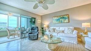 Ein Wohnzimmer mit Glastisch, grauem Sessel, weißem Sofa und Deckenventilator.
