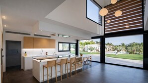 A modern kitchen with a central island, bar stools, and large windows offering a view of a landscaped outdoor area. - Luxury Villa West Nice (Nice)