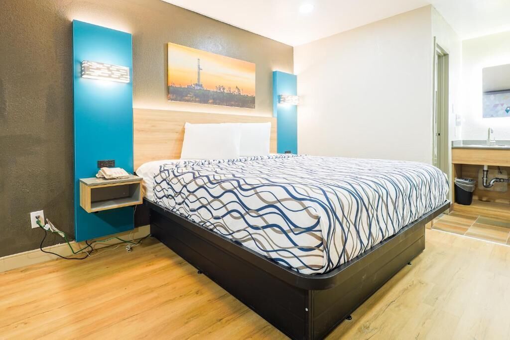 A modern hotel room with a large bed, a nightstand, a wall-mounted shelf, and a painting of a cityscape.