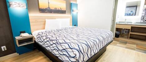 A modern hotel room with a bed, a nightstand, a painting, a bathroom with a mirror, and a sink.