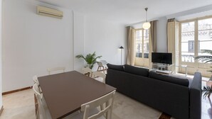 Un salon comprenant un canapé, une table à manger, des chaises, un téléviseur et une grande fenêtre avec des rideaux.