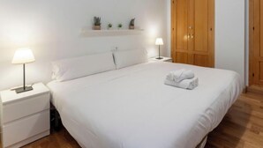 Un lit bien fait, avec du linge de lit blanc, deux lampes de chevet, une petite étagère avec des plantes en pot et une armoire en bois.