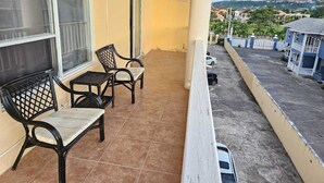 Un balcon avec deux chaises et une petite table, une fenêtre et une vue sur un quartier résidentiel avec des maisons et des voitures garées.