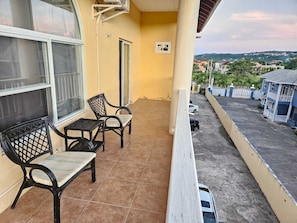 A balcony with two chairs and a table, a window, and a view of a residential area. - 2BR/2BA Coastal Escape Near Ocho Rios & Beaches (Tower Isle)