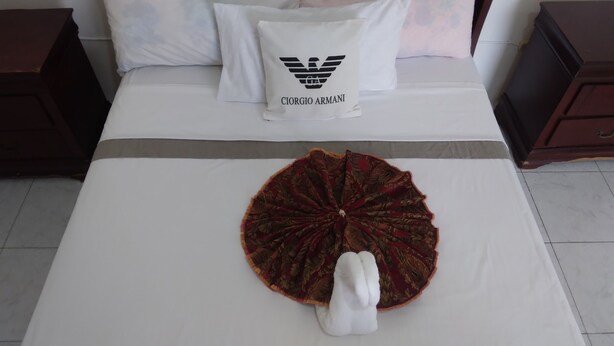 Un lit bien fait, avec un oreiller blanc orné d’un logo, un tissu décoratif rouge et or, et une serviette blanche.