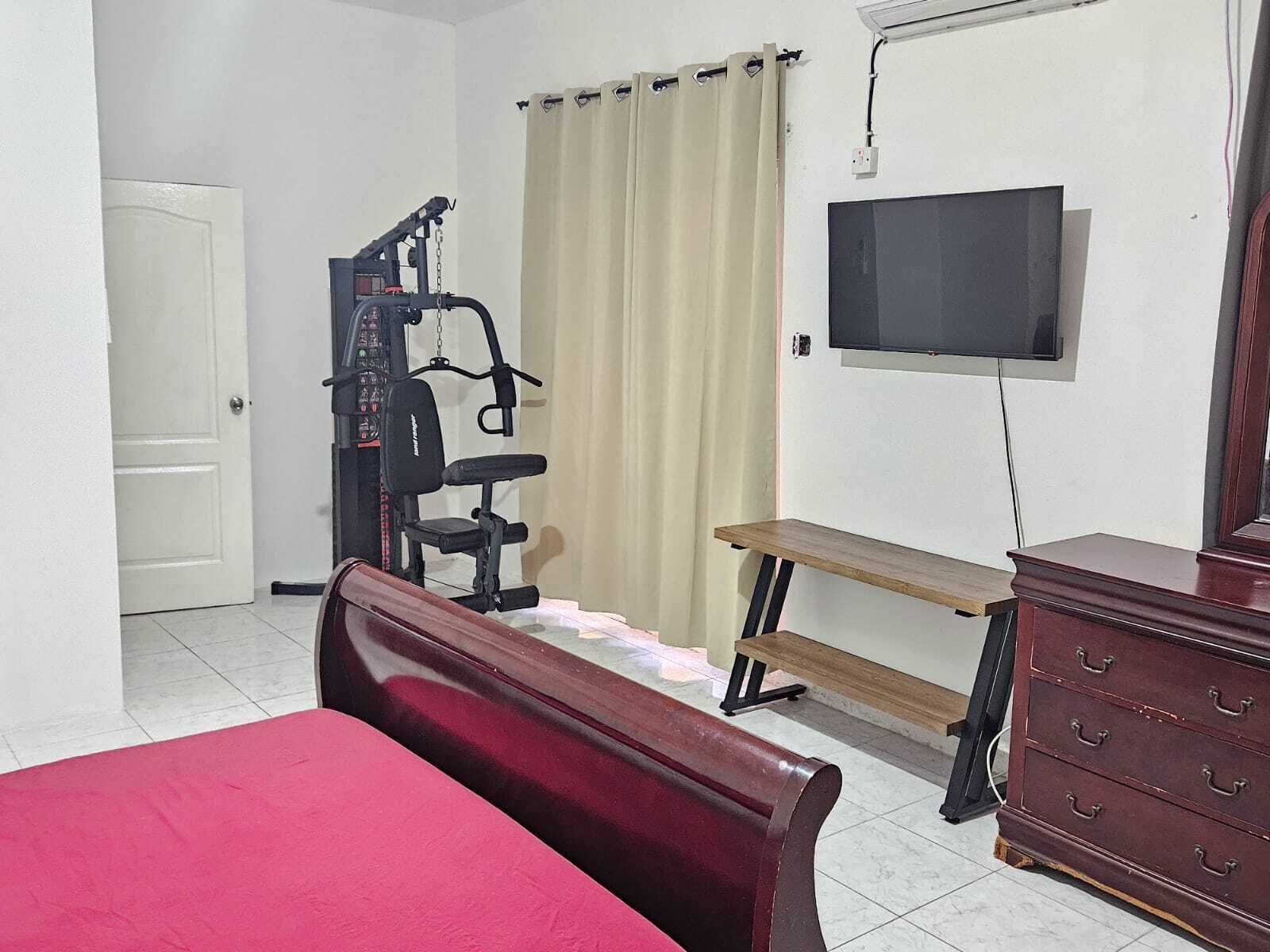 A bedroom with a bed, a desk, a chair, a TV, and a workout machine.