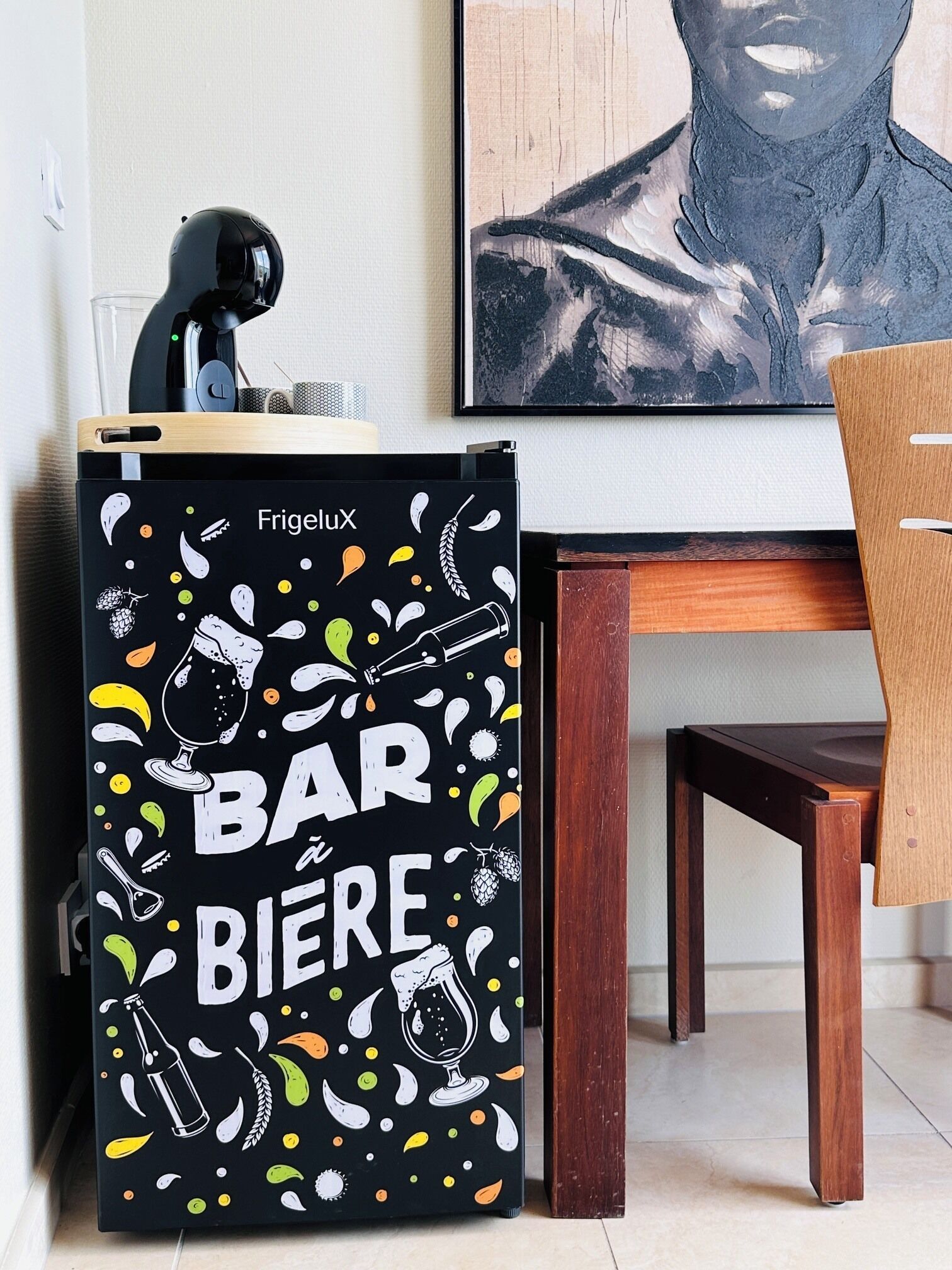 Un réfrigérateur noir avec les inscriptions « FrigluX » et « BAR BIÈRE », une chaise en bois et une table.