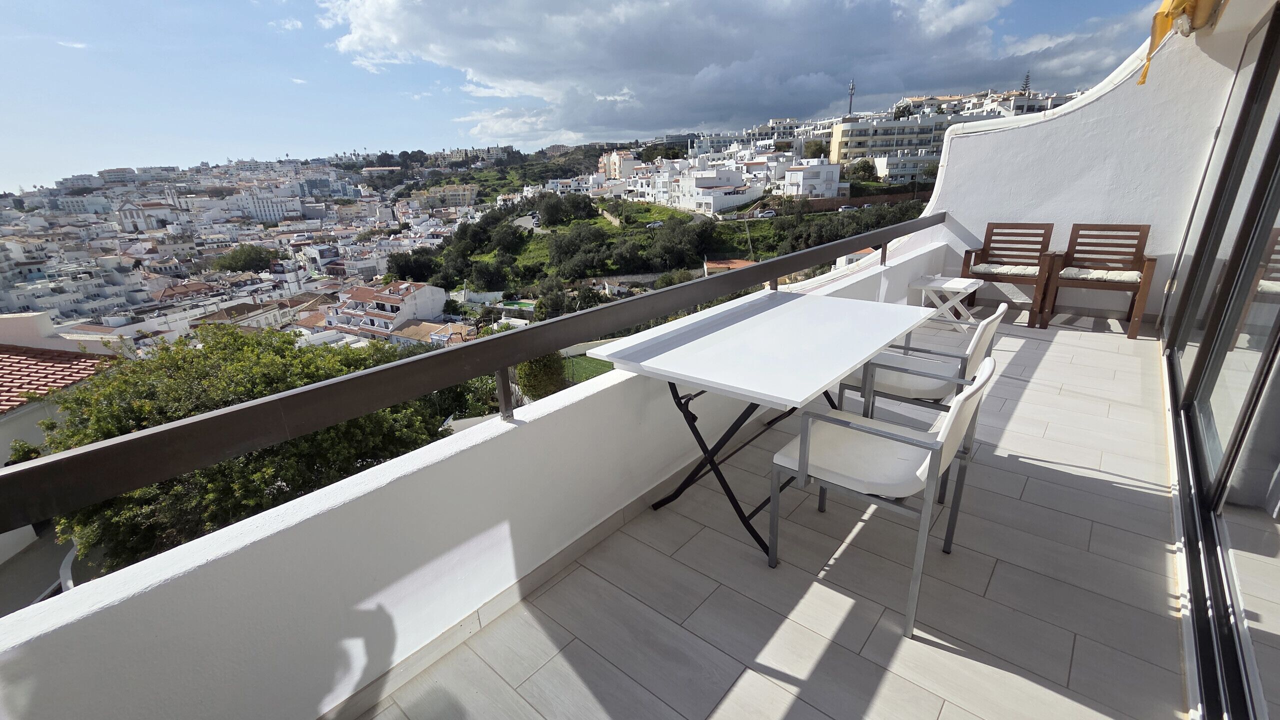 A balcony with a white table and chairs, overlooking a cityscape with buildings and greenery.