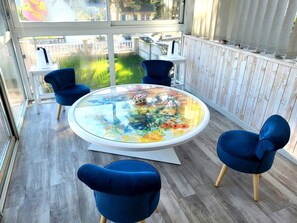 A modern room with a round glass table, blue chairs, and a view of a garden through large windows.