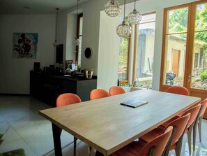 Een eetgedeelte met een houten tafel en oranje stoelen, een keuken met een zwart aanrecht en hangende lampen.