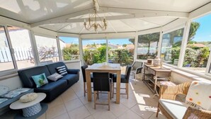 Eine überdachte Terrasse mit Tisch und Stühlen, einer Couch, Korbmöbeln und großen Fenstern mit Blick auf die Umgebung.