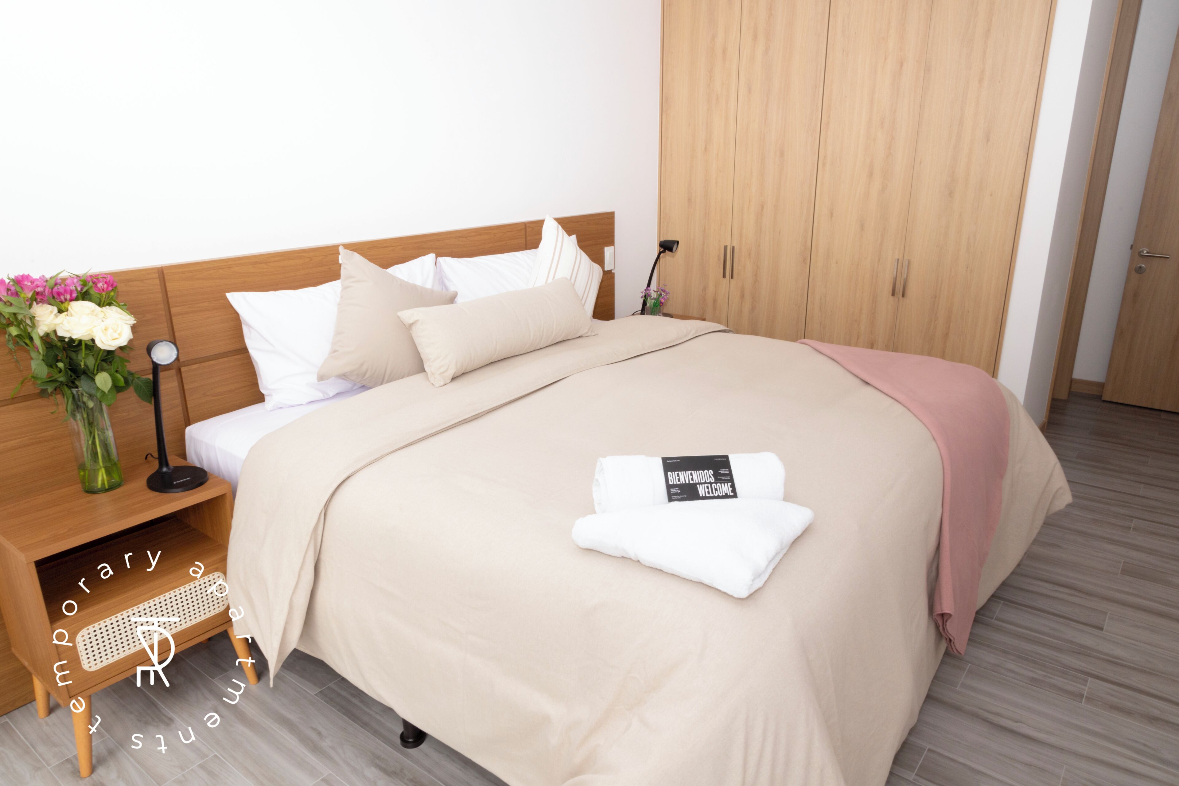 A neatly made bed with a beige bedspread, pillows, and a folded towel with a welcome sign. There is a wooden nightstand with a lamp and a vase of flowers. A wooden wardrobe is in the background.