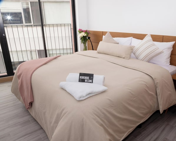 A neatly made bed with a welcome sign, a wooden headboard, and a view of a balcony with a railing. - Top Rentals EpiQ (Quito)