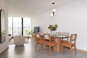 A modern dining area with a wooden table, four chairs, a vase with flowers, and a large window offering a city view.