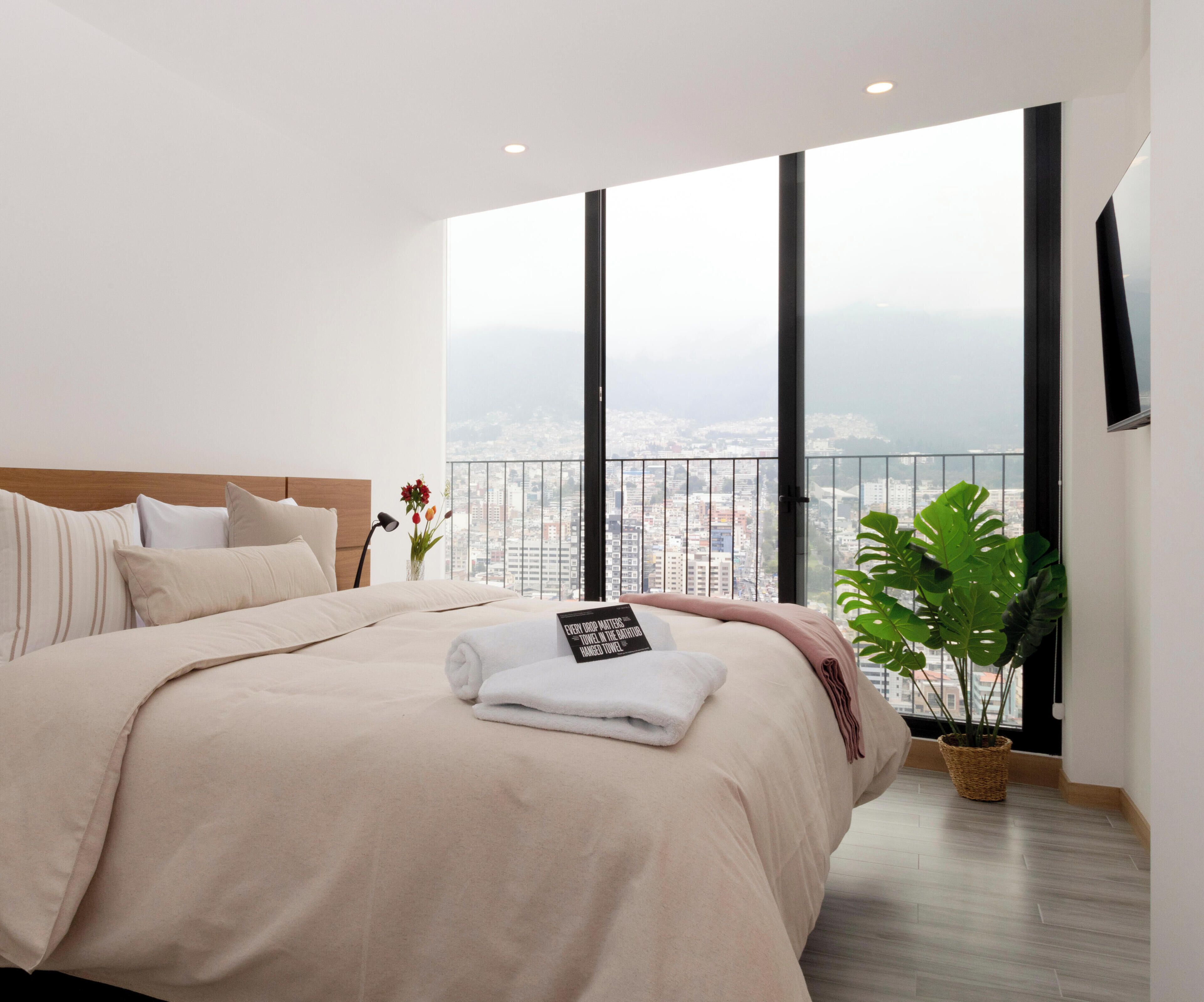 A modern bedroom with a large bed, a city view through floor-to-ceiling windows, and a potted plant.