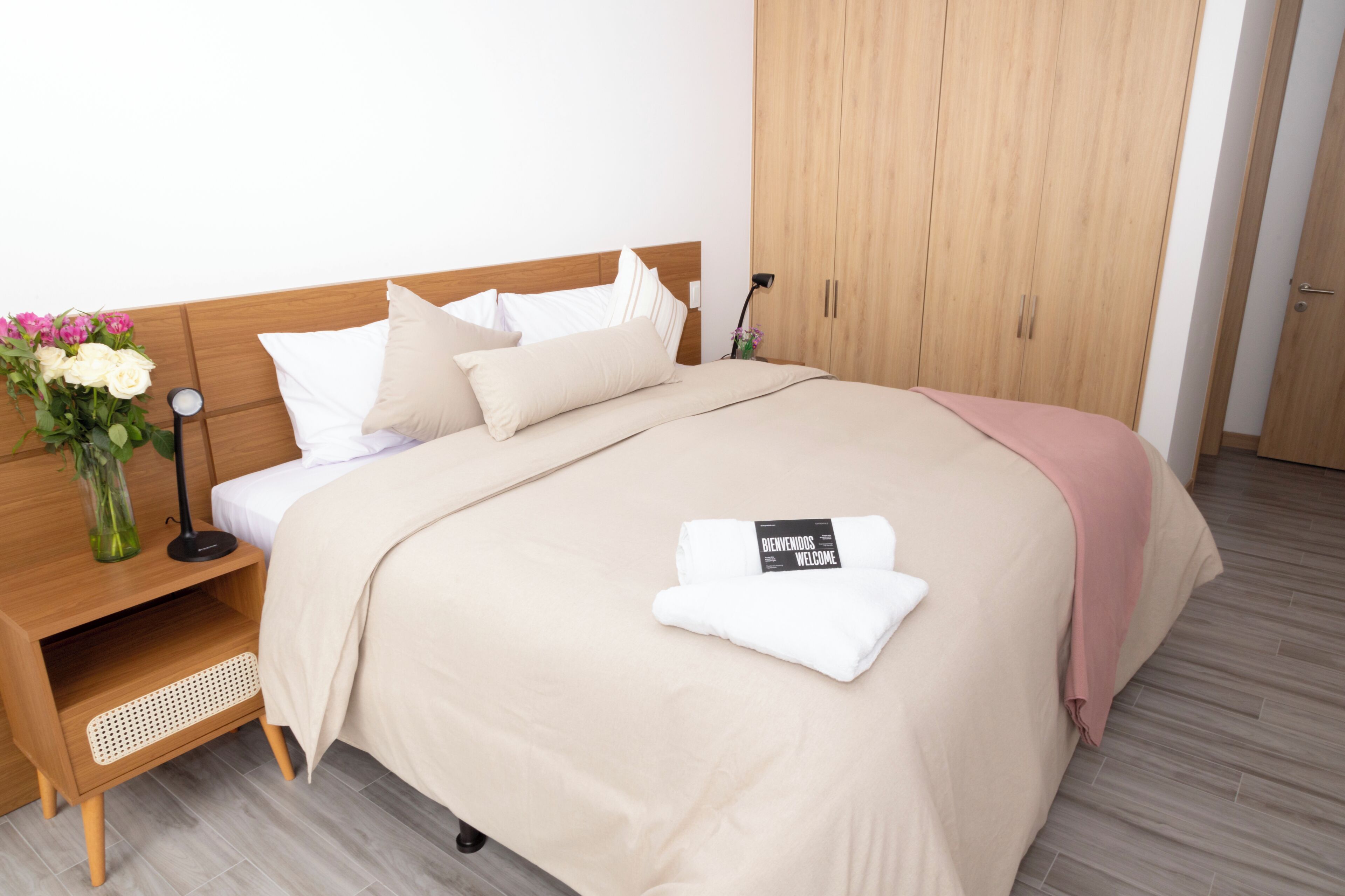 Un letto rifatto con cura, con copripiumino e cuscini color beige, un comodino con una lampada e un vaso di fiori, e un armadio in legno.