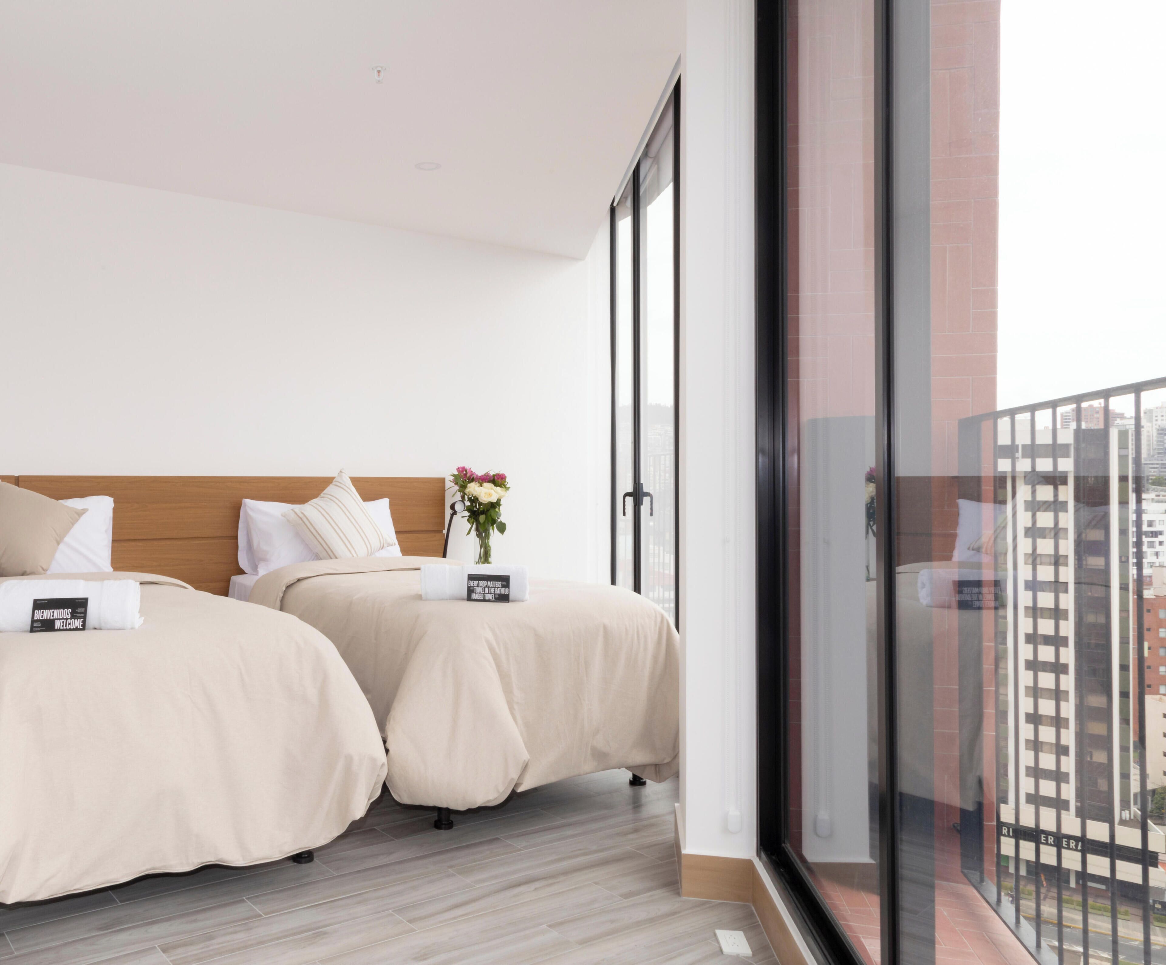 Ein Hotelzimmer mit zwei Betten, einem Nachttisch mit Blumenvase und einem großen Fenster mit Blick auf die Stadt.