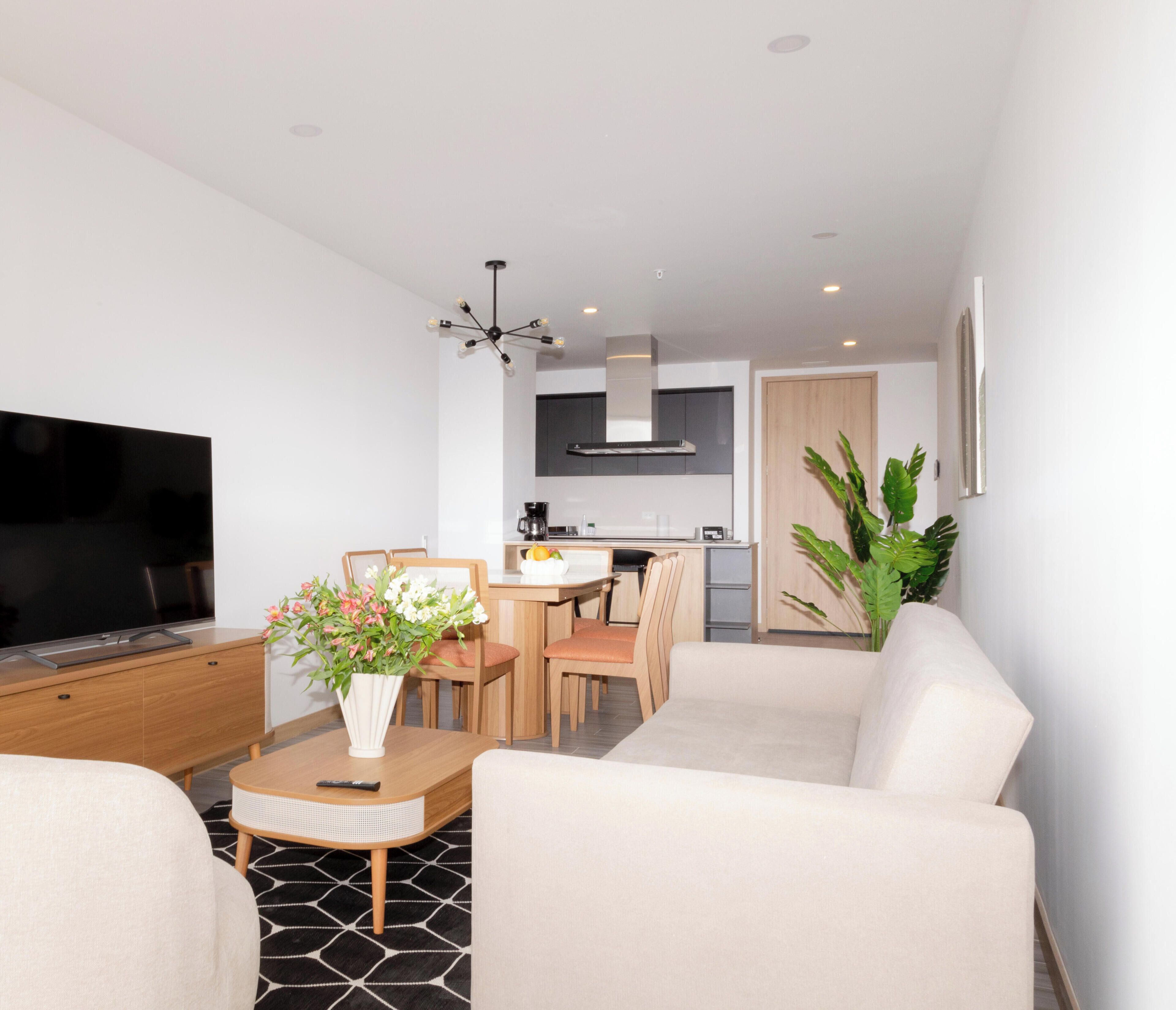 A modern living room with a flat-screen TV, a sofa, a coffee table with a vase of flowers, and a dining area with chairs and a table.