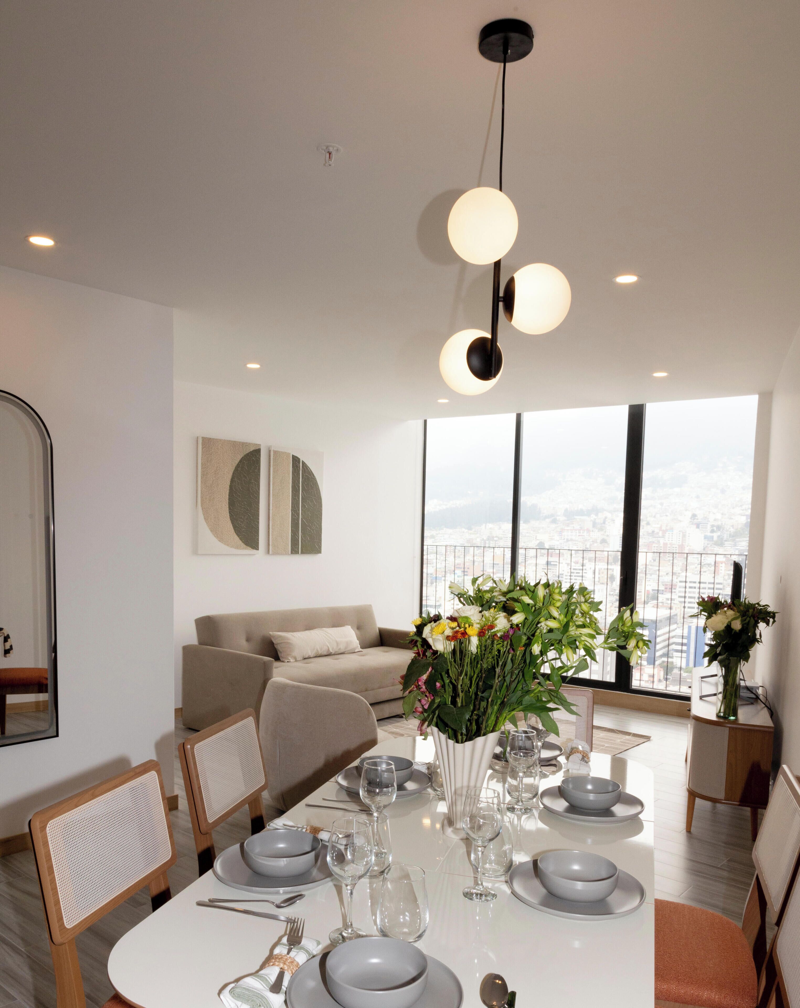 Un espace de repas moderne avec une table blanche dressée pour quatre personnes, un canapé et une vue sur la ville grâce à de grandes fenêtres.