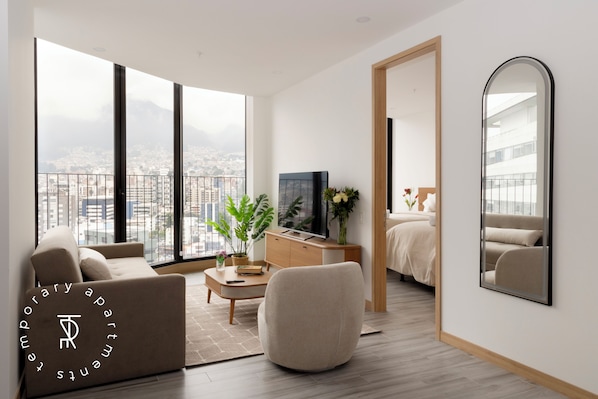 A modern living room with a large window, a sofa, a coffee table, a TV, and a bed. - Top Rentals EpiQ (Quito)